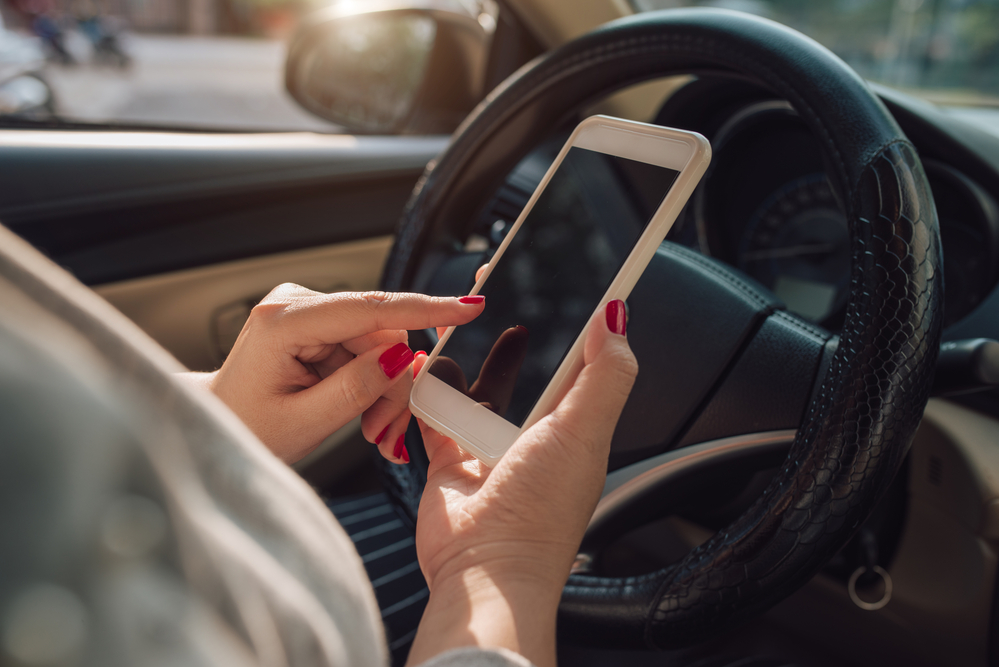 Ways You Can Minimize Distracted Driving - Mockup image of woman hand using mobile smartphone with blank screen while driving car and leaving home. clipping path.